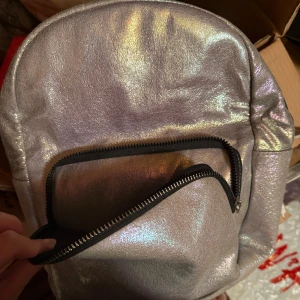 Silvrig ryggsäck från Primark - Cool ryggsäck i skimrande silver från Primark. Väskan har ett stort huvudfack, en praktisk ytterficka med dragkedja och justerbara axelband. Materialet är syntet med metallic-finish som ger en futuristisk vibe. Perfekt för dig som vill sticka ut i mängden.