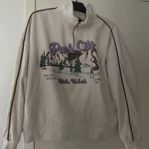 Vit half zip sweatshirt Park City - Vit sweatshirt med half zip och broderat Park City-tryck i lila och brunt framtill. Tröjan har långärmade ärmar med bruna ränder längs axlarna och en hög krage. Materialet är mjukt och känns som bomull eller polyester, perfekt för chill dagar. Katt finns i hemmet!
