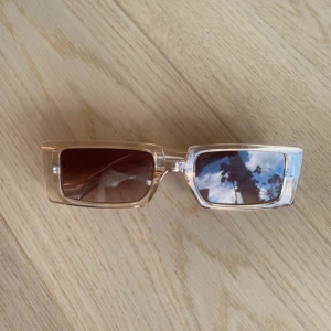 Rektangulära solglasögon i beige plast - Säljer ett par solglasögon i ordentligt och hållbart material, helt nya och aldrig använda. Perfekta för både vardag och semester, dessa solglasögon kombinerar stil och funktion. Passa på att göra ett kap och få solglasögon i toppskick!  Vänligen läs min bio innan du gör ett köp; där hittar du bland annat information om leveransprocessen med mera 😊  #solglasögon #sunglasses #accessoarer #sommarstil #summeressentials .