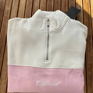 Vit och rosa half zip sweatshirt Official - Vit och ljusrosa sweatshirt från Official med halv dragkedja och hög krage. Tröjan har broderad logga på bröstet, breda rosa paneler över bröst och ärmar samt mjukt bomullsmaterial. Perfekt för en chill och trendig look.