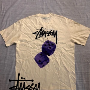 Stüssy t-shirt XL med tärningsprint - Gul t-shirt från Stüssy. svart logga på bröstet och stor grafisk print med lila tärningar på ryggen. Klassisk passform och rund hals. Perfekt för dig som gillar streetwear och vill sticka ut med en unik design.