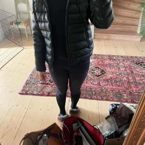 Svart Colmar pufferjacka med huva - Svart pufferjacka från Colmar Originals med glansig finish, huva och dragkedja framtill. Jackan har två sidofickor med dragkedja och ett Colmar-märke på ärmen. Perfekt för dig som gillar en clean och sportig look under kyliga dagar.