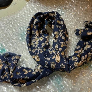 Blommig marinblå halsduk med fjärilar - Snygg marinblå halsduk med blommigt mönster och ljusa fjärilar i beige och vitt. Tunt och mjukt tyg, perfekt att slänga runt halsen för en chill look. Fransad kant ger extra vibe. Passar dig som gillar detaljer och vill sticka ut lite extra.