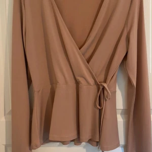 Beige omlottopp från Gina Tricot - Säljer en beige omlottopp från Gina Tricot i storlek M. Toppen har långa ärmar, v-ringad hals och knyt i midjan för en snygg siluett. Materialet är mjukt och stretchigt, perfekt för dig som gillar stilrena och enkla plagg med lite extra detalj.