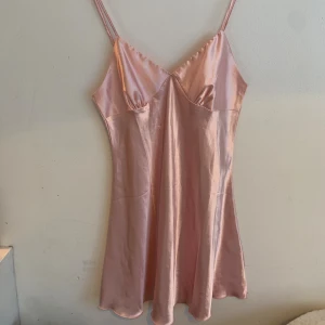 Ljusrosa satin klänning med smala axelband - Supersöt ljusrosa kortklänning i glansigt satinliknande material. Klänningen har smala justerbara axelband och v-ringad urringning. Den är figurnära upptill och faller snyggt nedtill. Perfekt för dig som gillar en enkel men elegant stil.