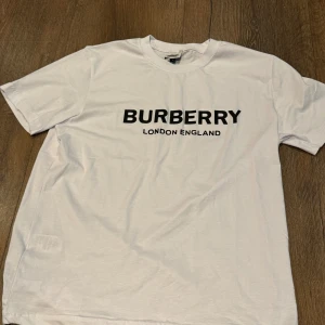 Vit Burberry t-shirt med tryck - Vit t-shirt från Burberry i storlek XL med klassiskt svart logotryck 'BURBERRY LONDON ENGLAND' på bröstet. Modellen har rund hals och korta ärmar, tillverkad i mjuk bomull för en skön känsla. Perfekt för dig som gillar stilrena designer och ikoniska märken. Passar både L och XL