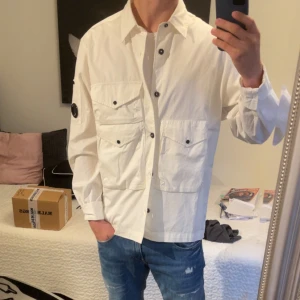 Vit overshirt från C.P. Company - Säljer en stilren vit overshirt från C.P. Company med klassisk krage, stora bröstfickor med knappar och den ikoniska linsen på ärmen. Skjortan är i bomull och har en clean look med svarta knappdetaljer. Overshirten har används ett då antal gånger.
