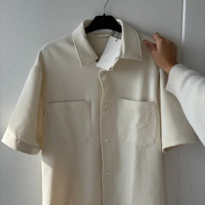 Beige kortärmad skjorta från Zara - Helt ny med prislapp och helt oanvänd.Stilren beige skjorta från Zara med korta ärmar, klassisk krage och två stora bröstfickor. Skjortan har knappar framtill och är tillverkad i ett mjukt bomullstyg. Perfekt för en clean och avslappnad look.