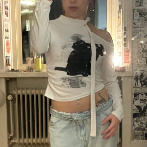 Vit offshoulder topp med kattmotiv - Säljer en vit långärmad topp med offshoulder-design och coolt svart kattmotiv på framsidan. Toppen har en unik knyt-detalj vid halsen och är köpt i sydkorea. passar xs-s