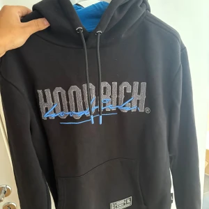 Svart/blå hoodie från Hoodrich  - Svart hoodie från Hoodrich med stor logga i grått och blått framtill och baktill. Tröjan har huva med snörning och en stor magficka. Mjuk insida och klassisk streetwear-look, perfekt för dig som gillar stilrena och bekväma plagg.