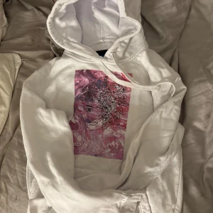 Vit hoodie med konstmotiv från The Cool Elephant - Vit hoodie från The Cool Elephant med en stor, färgstark konstnärlig print framtill. Motivet föreställer ett ansikte med blommor i håret i rosa, lila och röda toner. Tröjan har huva med snörning och är långärmad. Perfekt för dig som gillar unika prints men ändå en avslappnad look.🤍