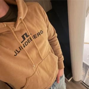 Beige hoodie från J.Lindeberg - Snygg beige hoodie från J.Lindeberg med svart logotyp på bröstet. Tröjan har huva med snörning, stor magficka och ribbade muddar. Mjuk insida i fleece och klassisk passform, perfekt för chill dagar. Passar dig som gillar stilrena och bekväma plagg.