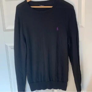 Svart merinoulltröja från Polo Ralph Lauren - Svart långärmad tröja från Polo Ralph Lauren i mjuk merinoull. Slim fit-modell med rund hals och lila broderad logga på bröstet. Perfekt för dig som gillar stilrena och klassiska plagg med en twist.