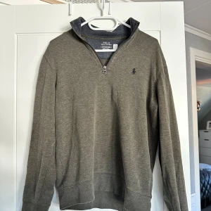 Olivgrön half zip tröja Polo Ralph Lauren - Olivgrön långärmad tröja från Polo Ralph Lauren med klassisk broderad logga på bröstet. Tröjan har en hög krage med half zip-dragkedja och är tillverkad i mjuk bomullsmix. Perfekt för lager-på-lager och chill stil.