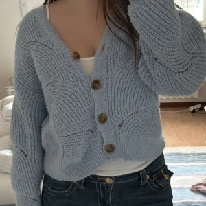 Ljusblå stickad kofta med träknappar - Superfin ljusblå stickad kofta med stora träknappar och snyggt mönster i garnet. Koftan har v-ringning, lång ärm och en lite croppad passform. Perfekt att slänga över ett linne eller t-shirt för en chill look.