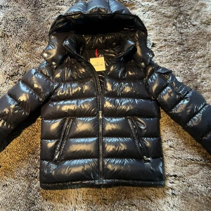  Moncler Maya  - Snygg svart pufferjacka från Moncler med glansig finish, huva och två dragkedjeförsedda fickor fram. Jackan har broderad Moncler-logga på ärmen och quiltad design. Perfekt för dig som vill ha en varm och stilren jacka med exklusiv känsla.