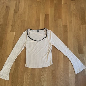 Vit ribbad långärmad topp från SHEIN - Säljer en vit ribbad långärmad topp från SHEIN med svarta kanter runt halsen. Toppen har utsvängda ärmar och ett smickrande v-ringad snitt. Perfekt att styla med jeans eller kjol för en trendig look.