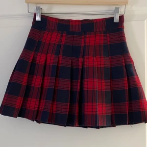 Rutig röd och mörkblå plisserad kjol - Supersnygg kort plisserad kjol i rött och mörkblått rutigt mönster från Fashion. Kjolen har bred linning och är helfodrad med svart innerfoder. Perfekt för dig som vill ha en klassisk men edgy stil.