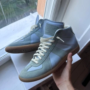 Maison Margiela blå sneakers i mocka och skinn - Säljer ett par stilrena Maison Margiela sneakers i ljusblått skinn och mocka med beige snörning och gummisula. Klassisk high-top modell med ikonisk Margiela-tunga och diskret branding. Perfekta för dig som gillar minimalistisk och clean stil.
