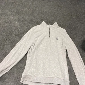 Ljusgrå långärmad tröja från U.S Polo ASSN. Ralph Lauren - Snygg ljusgrå långärmad tröja från Polo Ralph Lauren med half zip och liten broderad logga på bröstet. Perfekt för dig som gillar stilrena och bekväma plagg. Tröjan har en mjuk bomullskänsla och passar till många olika outfits.