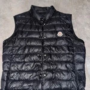 Svart dunväst från Moncler - Snygg svart dunväst från Moncler med quiltad design och klassisk logotyp på bröstet. Västen har tryckknappar framtill och hög krage som ger extra stilpoäng. Perfekt att slänga över en hoodie eller tröja för en cool look.