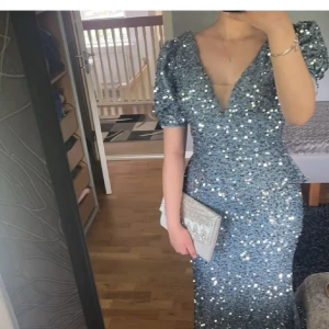 Glittrig långklänning med rosett - Säljer en fantastisk glittrig långklänning i ljusblå med v-ringning och puffärmar. Den har öppen rygg och en jätte fin stor rosett. Klänningen har figurnära passform och är täckt av paljetter som ger en riktigt festlig look. Perfekt för bal eller festkväll!