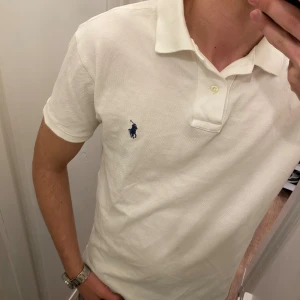 Vit piké från Polo Ralph Lauren (Slim Fit) - Vit pikétröja från Polo Ralph Lauren med klassisk krage och två knappar framtill. Tröjan har korta ärmar och den ikoniska blå broderade loggan på bröstet. Tillverkad i mjuk bomull som är skön mot huden och har en normal passform. Liten i storleken mellan small och Medium. Snabb Leverans!