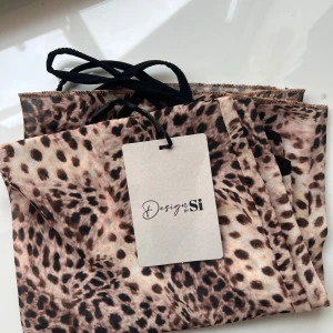 Leopardmönstrad klänning Design by Si - Snygg kort klänning från Design by Si med leopardmönster i beige och bruna toner. Klänningen har tunna axelband och v-ringad halsringning med svarta detaljer. Lite genomskinlig men perfekt till stranden eller styla. Längd=62cm Midja=34cm Byst=32cm storlek XS/S Aldrig använd