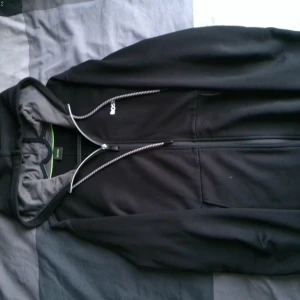 Svart hoodie från BOSS med dragkedja - Svart hoodie från BOSS med hel dragkedja framtill, snörning i huvan och grått innerfoder. Klassisk och stilren design med BOSS-logga på bröstet. Perfekt för chill dagar eller när du vill ha en clean look.