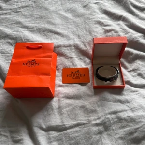 Svart armband från H - Säljer ett stilrent svart armband från H med den ikoniska H-detaljen i silverfärgad metall. Kommer med original ask, påse och certifikat. Perfekt accessoar för dig som gillar lyxiga och klassiska smycken med modern touch.