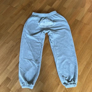 Grå mjukisbyxor från corteiz med tryck, M - Grå mjukisbyxor i bomull med elastisk midja och muddar vid benslut. Snyggt vitt tryck med motiv på baksidan. Byxorna har sidfickor och en bakficka med knapp. Perfekta för chill eller träning.