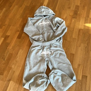 Grå mjukisbyxoroch hoodie sett från corteiz med tryck - Säljer ett par grå mjukisbyxor med elastisk midja och muddar vid bensluten. Byxorna har ett stort vitt tryck med motiv framtill, matchande med hoodie. Perfekt för chill dagar och riktigt skön passform.