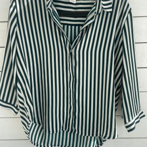 Randig skjorta från Monki i M - Snygg skjorta från Monki med vertikala gröna och vita ränder. Modellen har krage, knappar framtill och trekvartsärm. Skjortan är i ett mjukt och lätt polyestermaterial som känns skönt mot huden. Perfekt för dig som gillar en chill och trendig look.😇