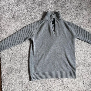 Grå stickad tröja med half zip - Grå stickad tröja från Peak Performance med ribbade ärmar och hög krage med half zip. Liten broderad logga på bröstet. Perfekt för kyliga dagar och enkel att matcha! Skriv vid minsta lilla fundering!