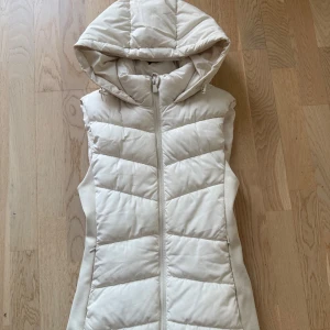 Beige dunväst med huva från Zara - Snygg beige dunväst från Zara med dragkedja framtill och stor, mysig huva. Västen har quiltad design och figurnära passform som ger en clean och modern look. Perfekt att slänga över en hoodie eller långärmad tröja när det är kyligt ute.