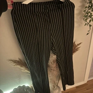 Svarta randiga kostymbyxor från Bubbleroom - Svarta kostymbyxor från Bubbleroom med vita smala ränder. Byxorna har raka ben och stängs med knapp och dragkedja. Klassisk look med midjehällor och diskret passform, perfekta för en stilren outfit.