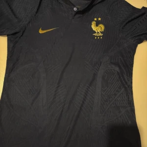 Frankrike Nike svart fotbollströja S - Svart fotbollströja från Nike för Frankrikes landslag med guldiga detaljer, broderad tupp och två stjärnor på bröstet. Tröjan har korta ärmar, diskret mönster i tyget och är tillverkad i Dri-Fit-material som andas.