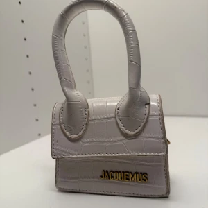 Vit väska Jacquemus - Superfin liten handväska från Jacquemus i vit, glansig skinnimitation med krokopräglat mönster. Väskan har två rundade handtag och guldiga detaljer, inklusive logotypen framtill. Perfekt för dig som gillar trendiga accessoarer med lyxig känsla.