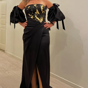 Svart långklänning med guldblommor - Elegant svart långklänning i satin med offshoulder och puffiga ärmar med knytband. Livet är dekorerat med guldiga blommor och pärldetaljer. Omlott-design med slits framtill ger en snygg siluett. Perfekt för dig som vill sticka ut på bal eller fest.