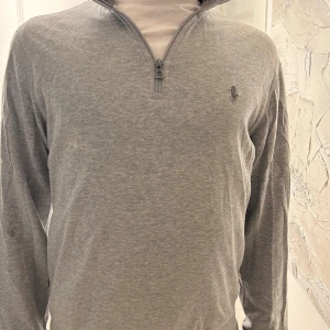 Grå half zip tröja från Polo Ralph Lauren - Snygg grå långärmad tröja från Polo Ralph Lauren med half zip och klassisk broderad logga på bröstet. Inga deffekter förutom en liten rosa fläck under armen, svår att se! Unik tröja, knappast använd. Kom dm för mer frågor