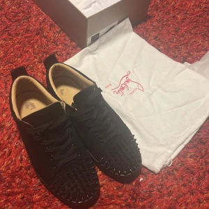 Svarta Louboutin sneakers med nitar - Säljer ett par svarta sneakers från Christian Louboutin med klassisk röd sula och coola svarta nitar på tån. Skorna har snörning och är tillverkade i mocka med detaljer i skinn. Kommer med originalkartong och dustbag.