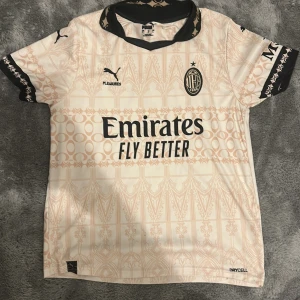 Äkta AC Milan x Puma kortärmad fotbollströja - Säljer en snygg AC Milan fotbollströja från Puma som är äkta den är ljus beige med svarta detaljer och grafiskt mönster. Tröjan har korta ärmar, svart krage och tryck från Emirates, Pleasures och wefox. Materialet är lätt och andas, perfekt för match eller träning.