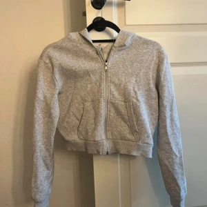 Grå croppad hoodie från H&M 146cm - Grå croppad hoodie från H&M med dragkedja framtill och huva. Tröjan har långa ärmar och en stor ficka på magen. Perfekt för chill dagar eller när du vill ha en enkel och stilren look.