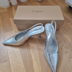 Silvriga pumps från Simmi London - Klackhöjd: 8cm ish. Glansiga pumps från Simmi London i metallic silver med spetsig tå och smal klack. Skorna har slingback-rem och ett diskret ormskinnsmönster.