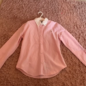 Rosa skjorta från Ralph Lauren XL - Snygg rosa skjorta från Ralph Lauren i 100% bomull. Klassisk modell med vit krage, knappar framtill och broderad logga på bröstet. Perfekt för dig som gillar stilrena och tidlösa plagg med en preppy vibe.