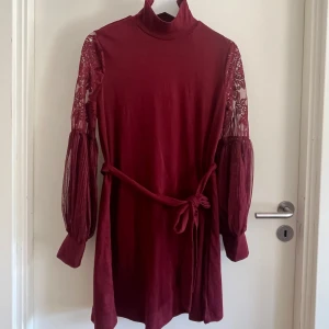 Vinröd klänning med spetsärmar  - Vinröd kortklänning från SHEIN med höghals, långa spetsärmar och transparenta detaljer. Klänningen har ett avtagbart bälte i midjan och ballongärmar med spets och mesh. 