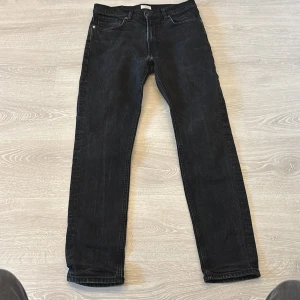 Svarta Taktil William Tapered jeans - Snygga svarta jeans från Taktil, modell William Tapered. De har en klassisk femficksdesign, raka ben med avsmalnande passform och diskret läderpatch bak i midjan. Perfekta för dig som gillar stilrena och bekväma jeans med modern siluett.