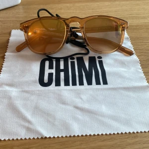 Bruna solglasögon från CHIMI - Snygga solglasögon från CHIMI med transparent brun båge och matchande bruna glas. Klassisk rundad form och cool retrostil. Kommer med svart glasögonband, putsduk och originaletui. Perfekta för att lyfta din look i solen!