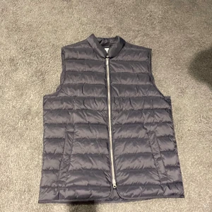 Mörkgrå quiltad dunväst med dragkedja - Snygg mörkgrå dunväst med horisontella quiltade sömmar och hel dragkedja framtill. Västen har en stilren, rak passform och två sidofickor. Perfekt att slänga över en hoodie för extra värme under kyliga dagar.