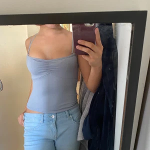 Ljusblå linnetopp med smala axelband - Säljer en ljusblå linnetopp med smala axelband och rynkdetalj framtill. Toppen är figurnära och har en enkel, stilren design som passar perfekt till jeans eller kjol. Perfekt för varma dagar eller lager på lager.